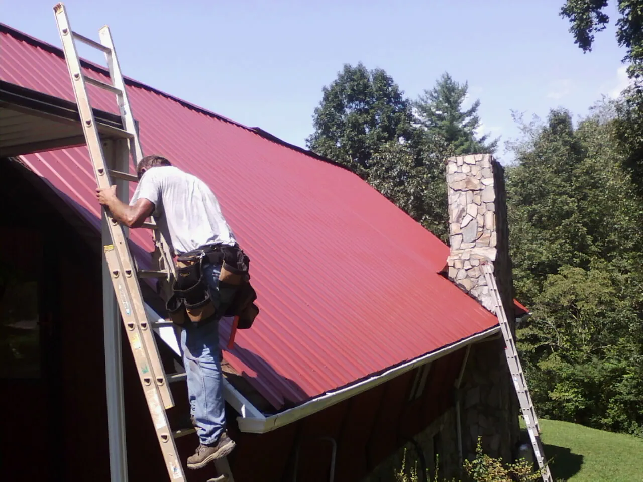 Roofer on a ladder beside a red metal roof and stone chimney — Soffit & Fascia Repair by Roofing Brothers in Paeonian Springs